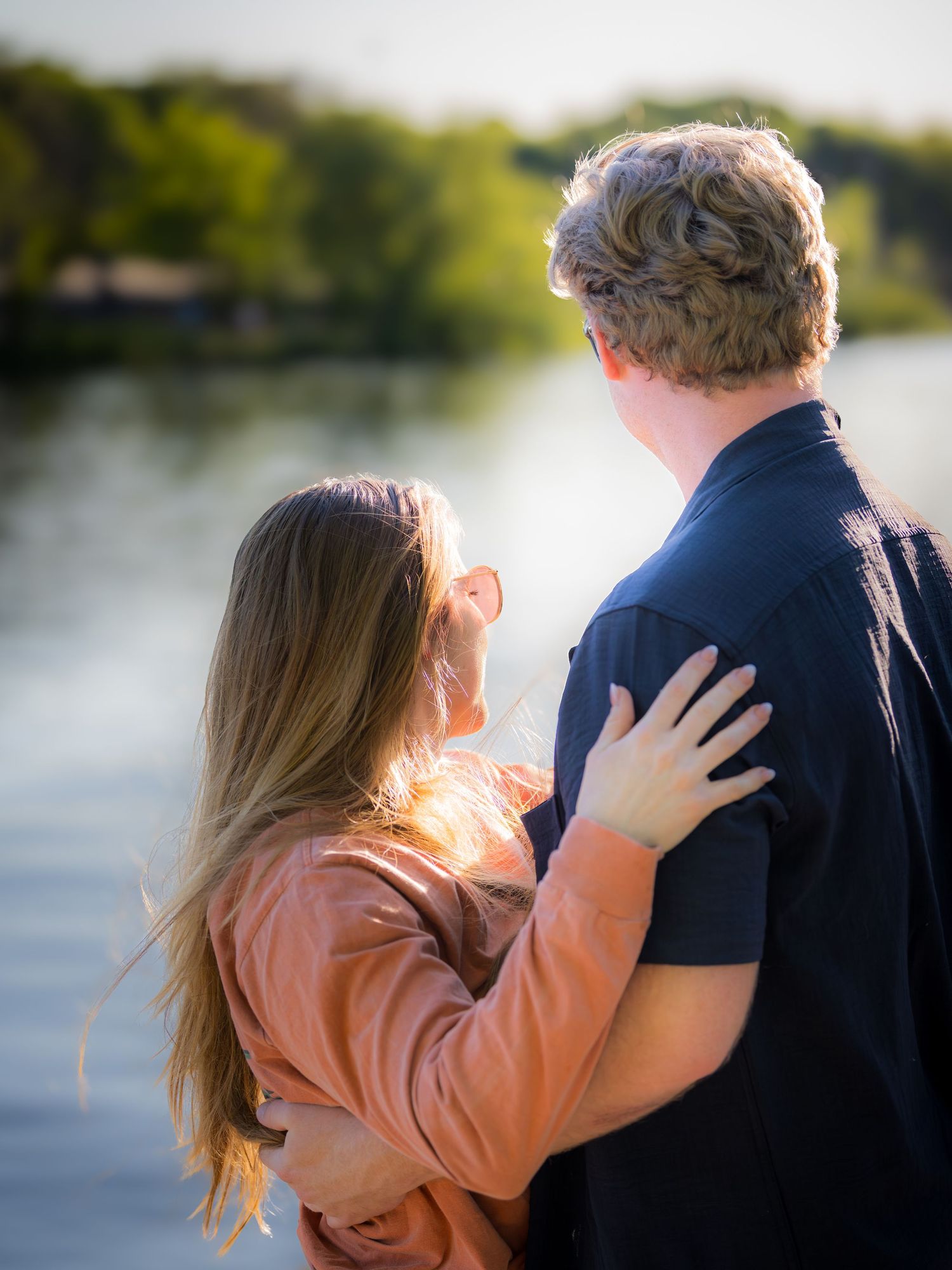 Couple embracing by a serene lake at golden hour, viewed from behind.