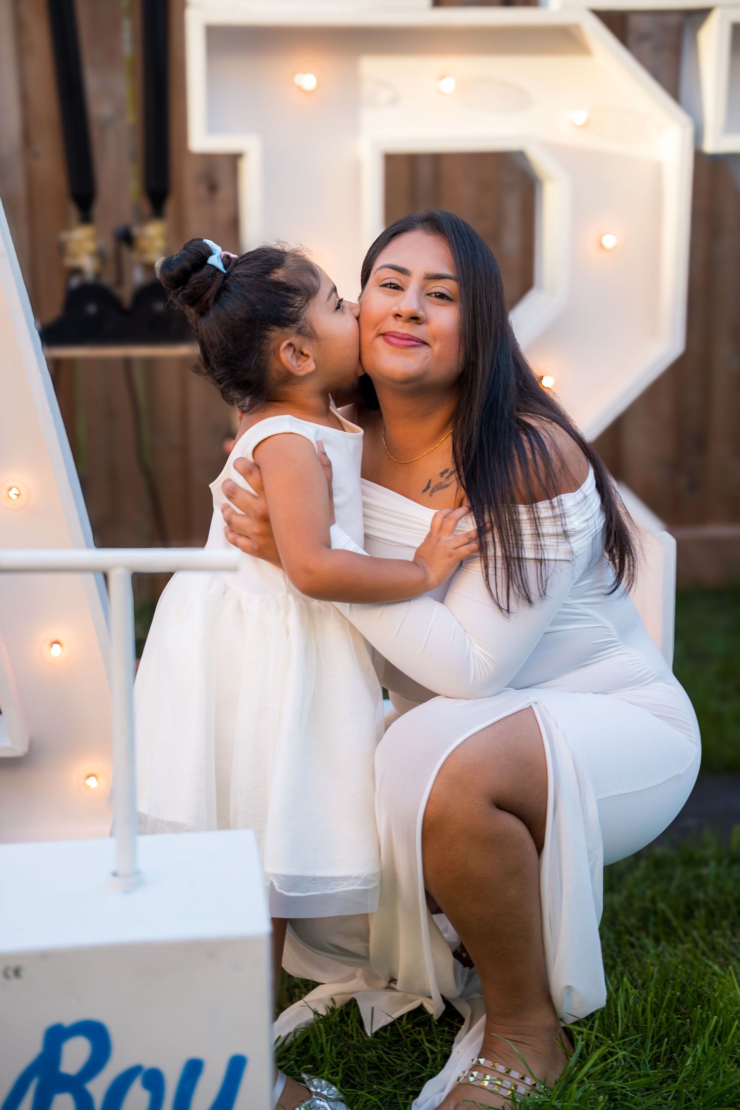 Young girl in white dress kisses mother's cheek at outdoor gender reveal party with lit letter decorations.