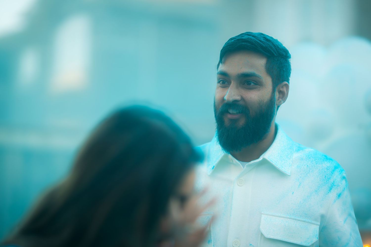 Bearded man in white shirt smiling during a blue powder gender reveal celebration outdoors.