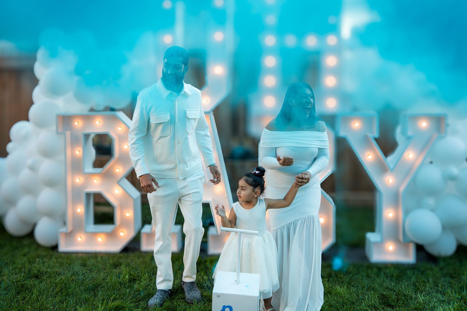 A family reveals baby gender with blue smoke beside a lit BABY sign and white balloons.