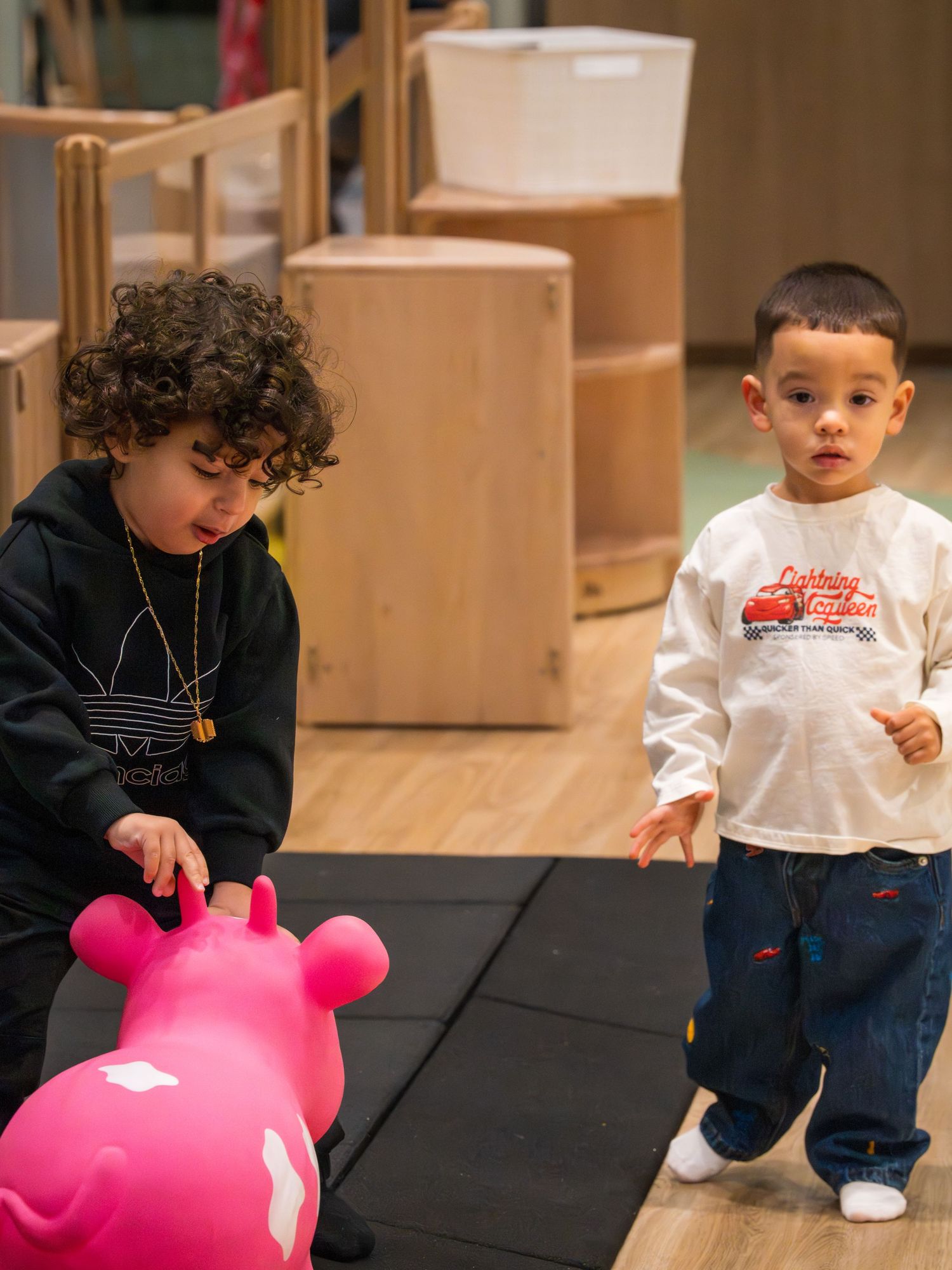 Two toddlers playing with a pink bouncy toy in a wooden play area.