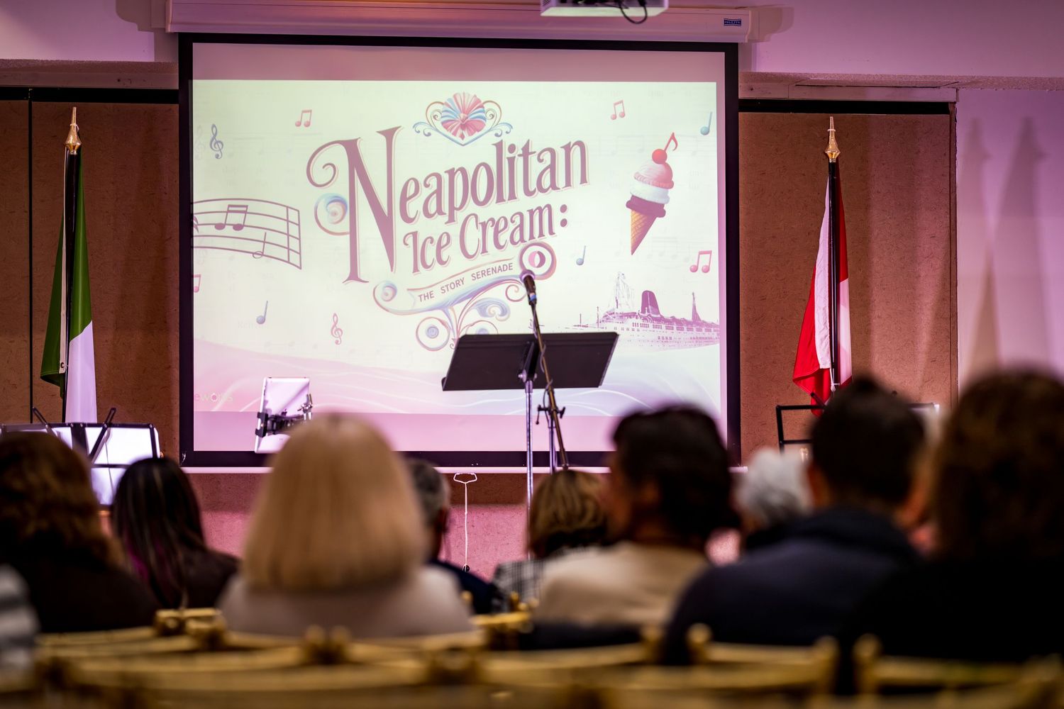Audience watches a presentation for Neapolitan Ice Cream musical projected on a large screen at an event.