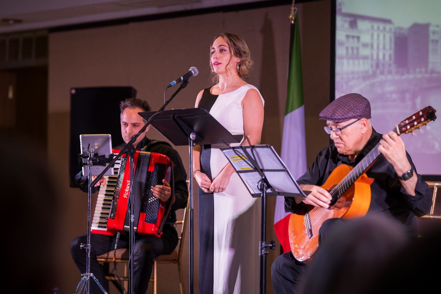 A female vocalist in a white dress performs on stage with an accordionist and guitarist at an Italian-themed event.
