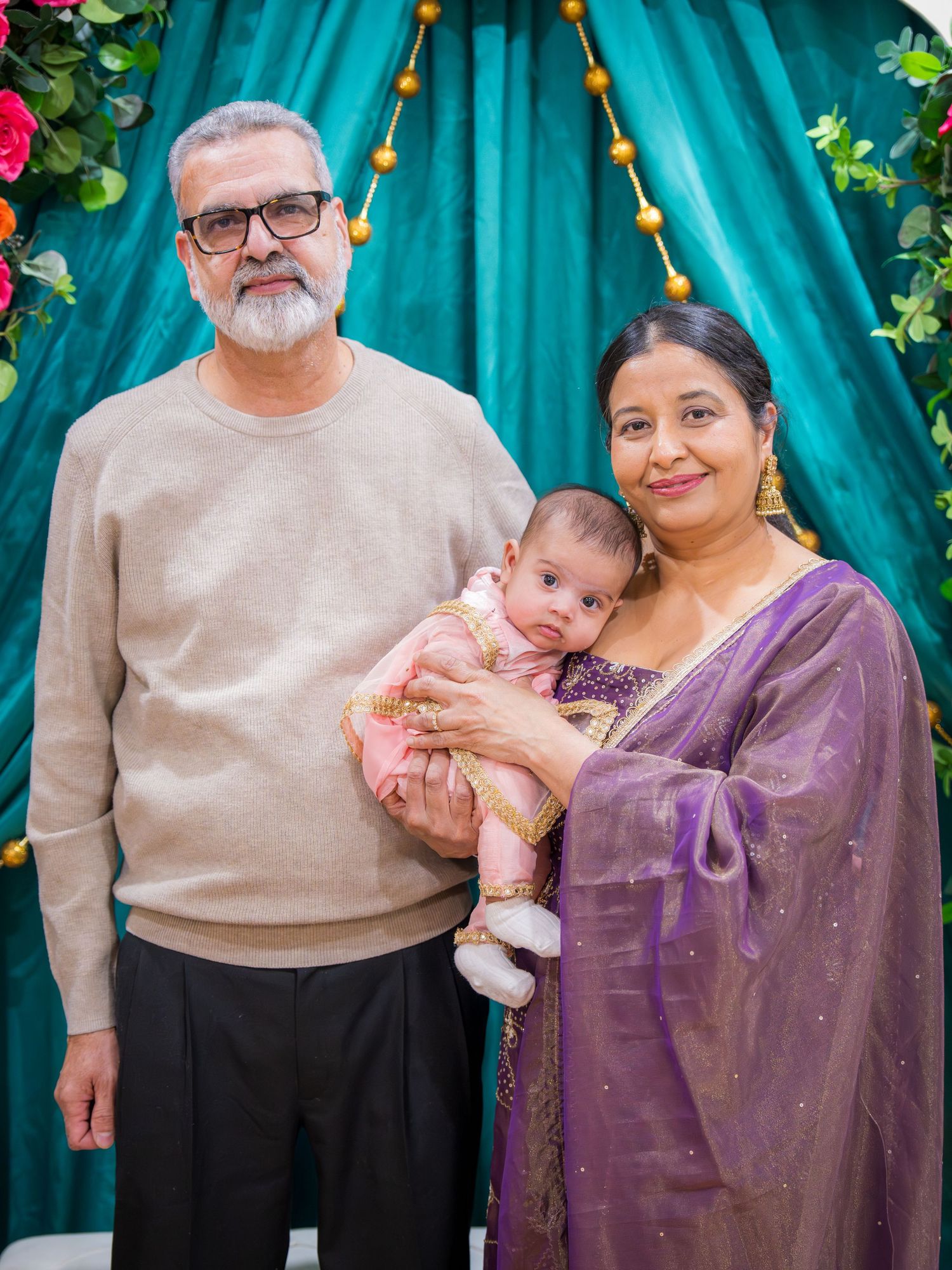 Couple in traditional South Asian attire holding newborn baby before teal floral decorated backdrop.