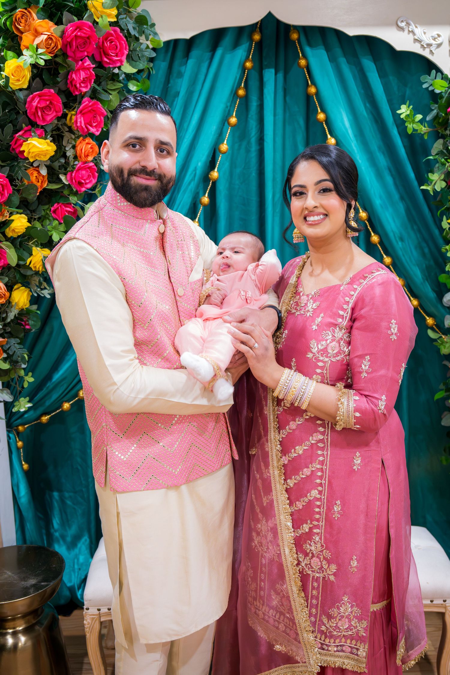 Indian couple in pink traditional attire holding newborn baby at decorated cultural ceremony.