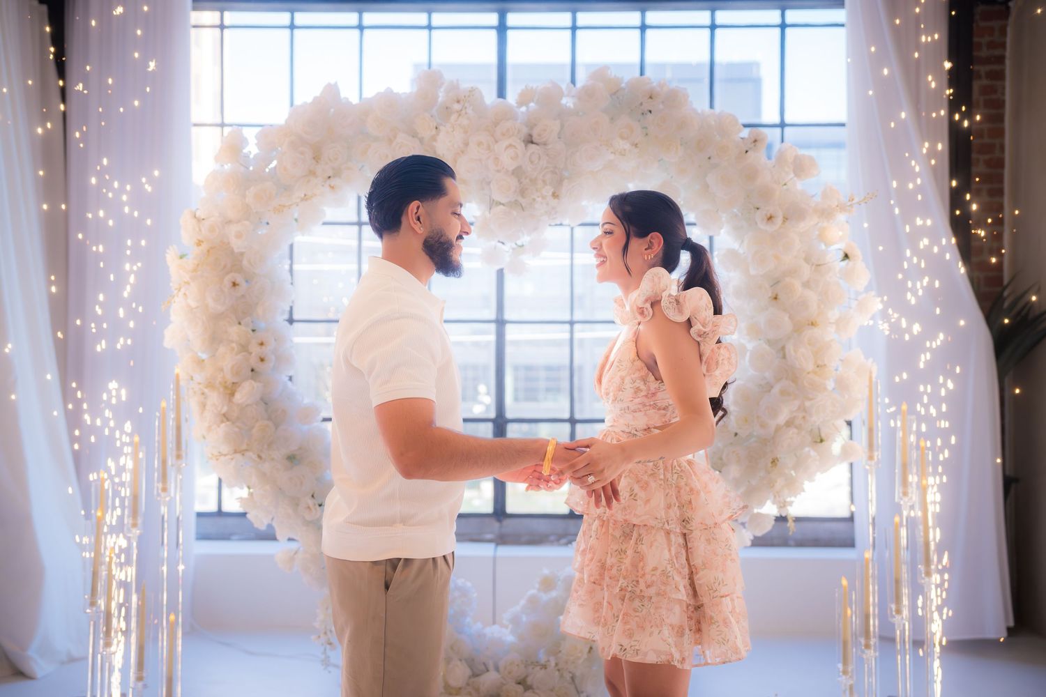 Couple holding hands and smiling before a stunning white floral heart arch with glowing fairy lights.