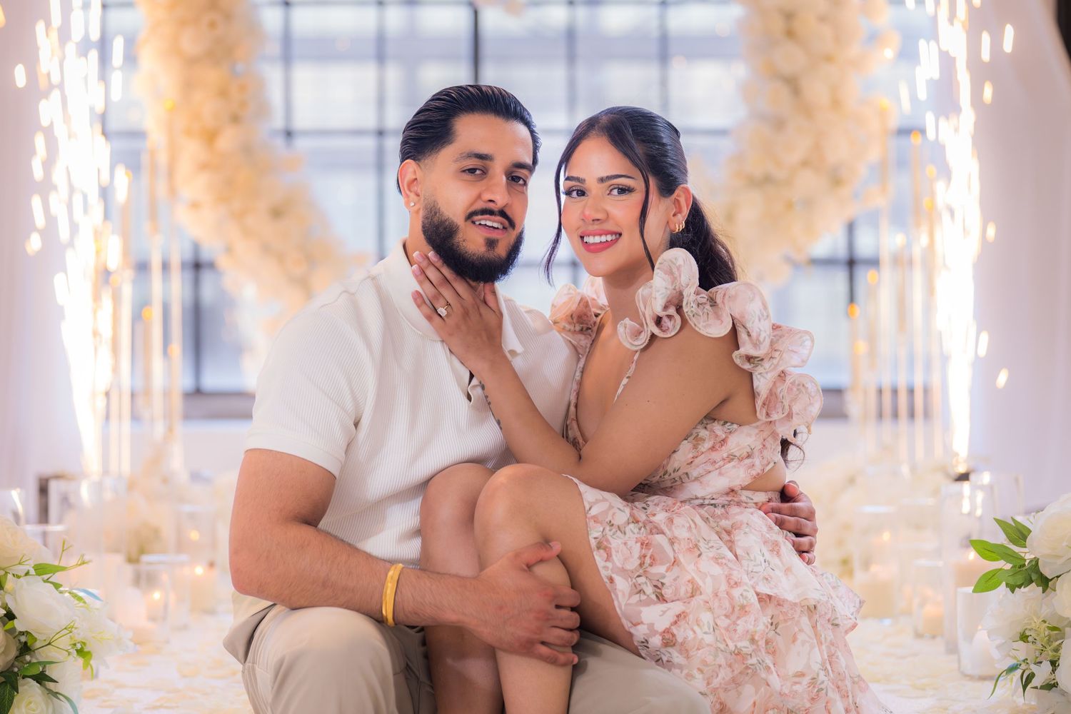 Smiling engaged couple sitting together surrounded by white floral arrangements and candles.