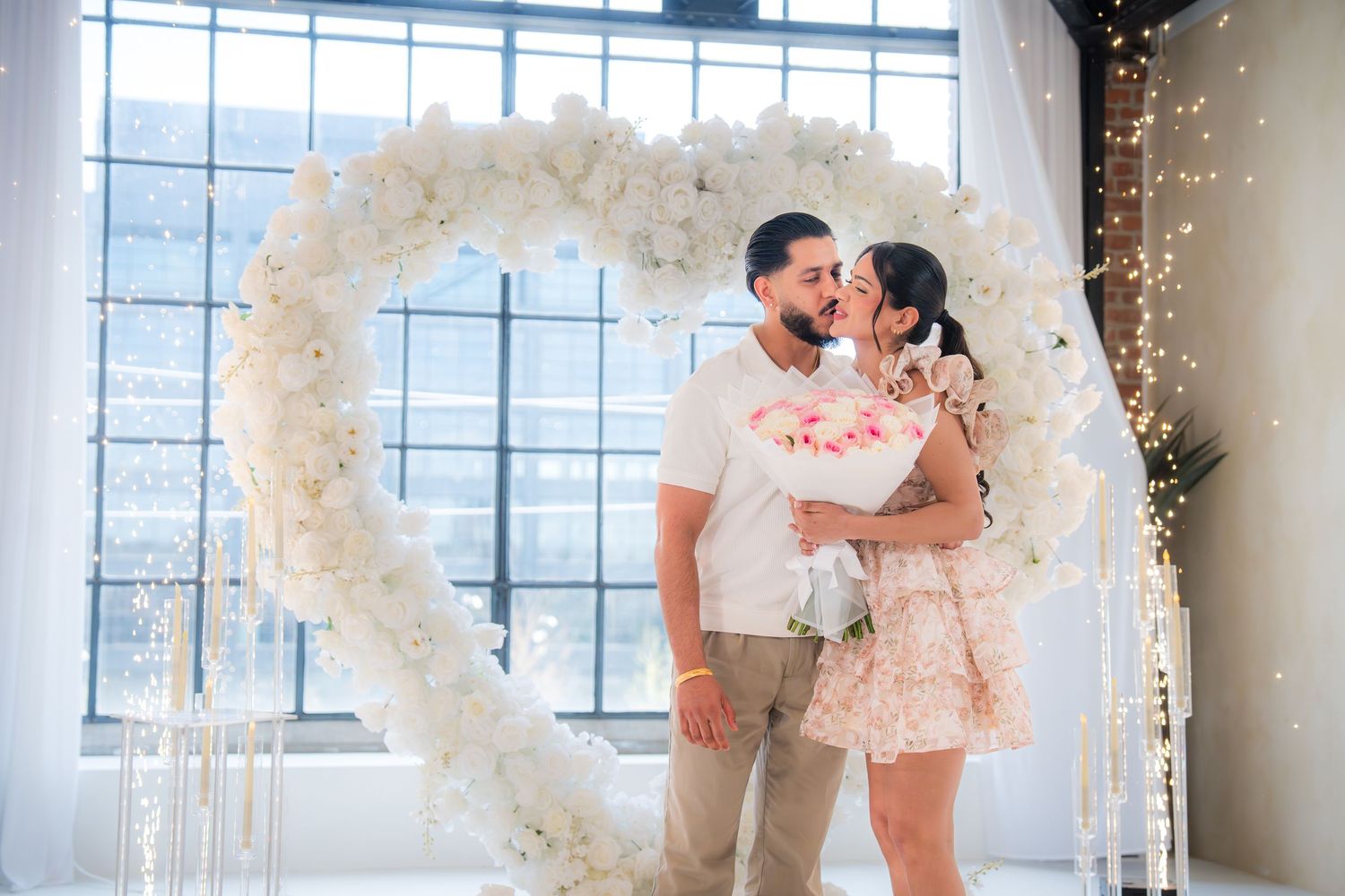 Couple embraces under a white floral heart arch with fairy lights during a romantic proposal setup.
