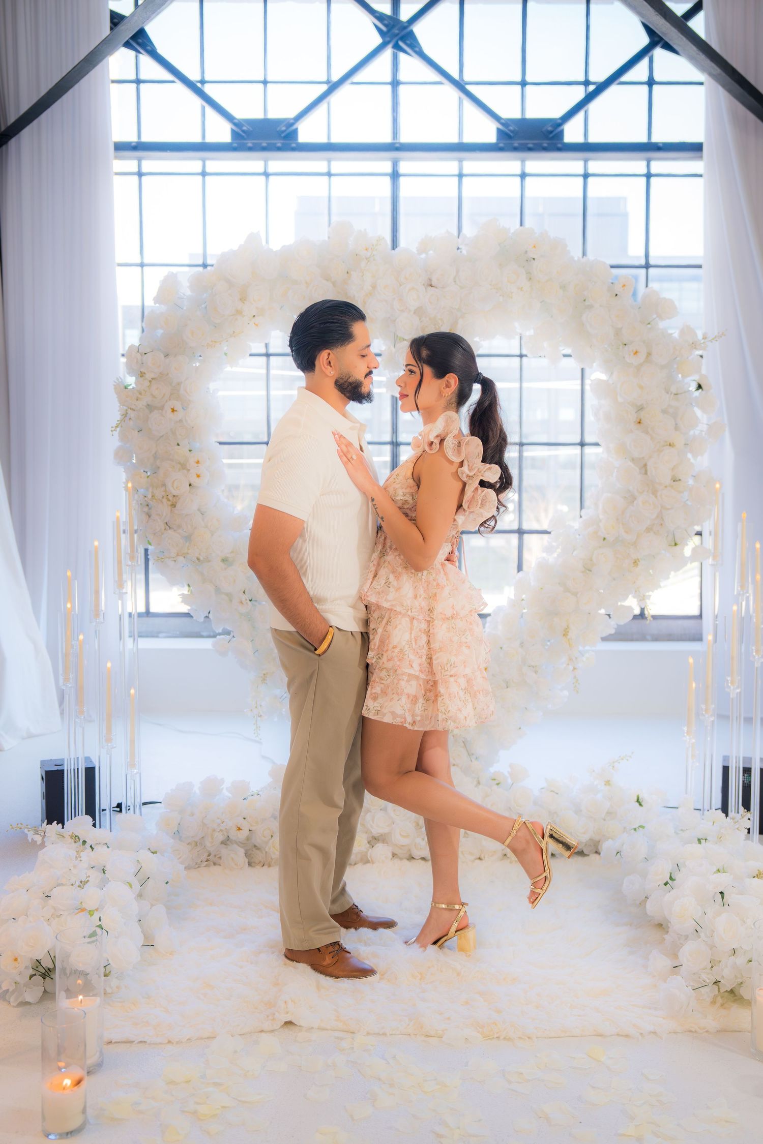 Couple embraces in front of a large white floral heart arch with candles and fairy lights.