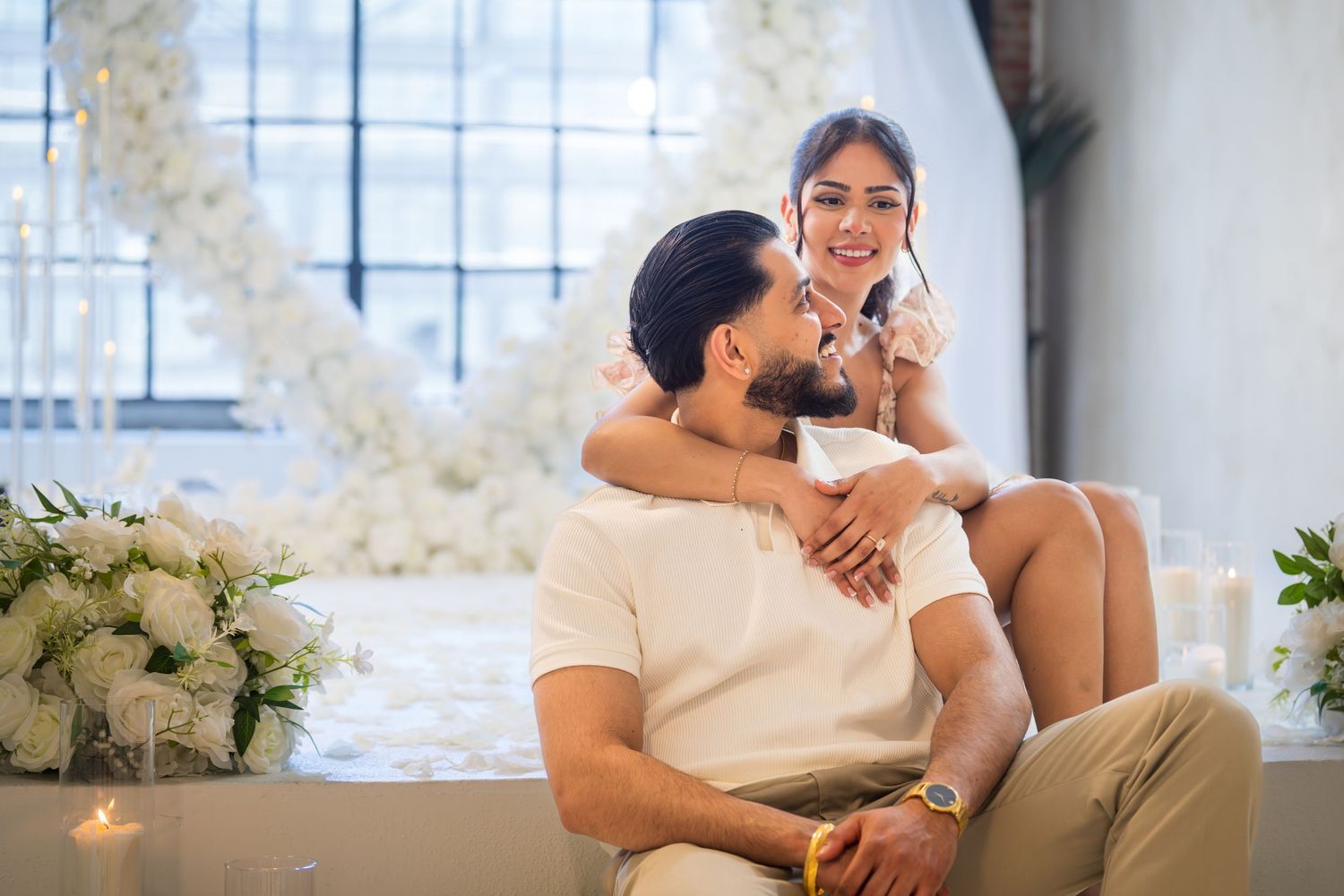 Couple sitting together smiling in front of white floral heart arch with candles and flowers.