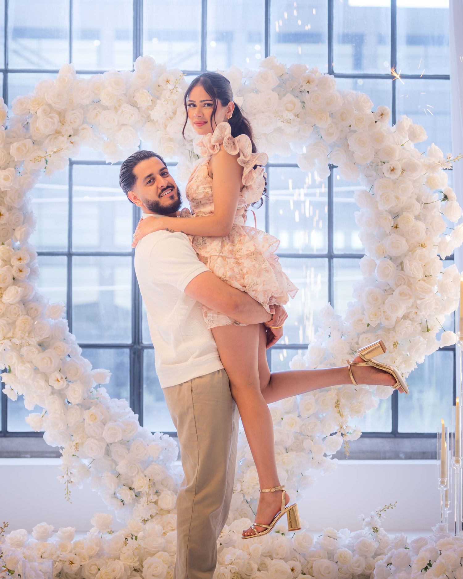 Newly engaged couple posing in front of large white floral heart arch, man lifting woman in gold heels.