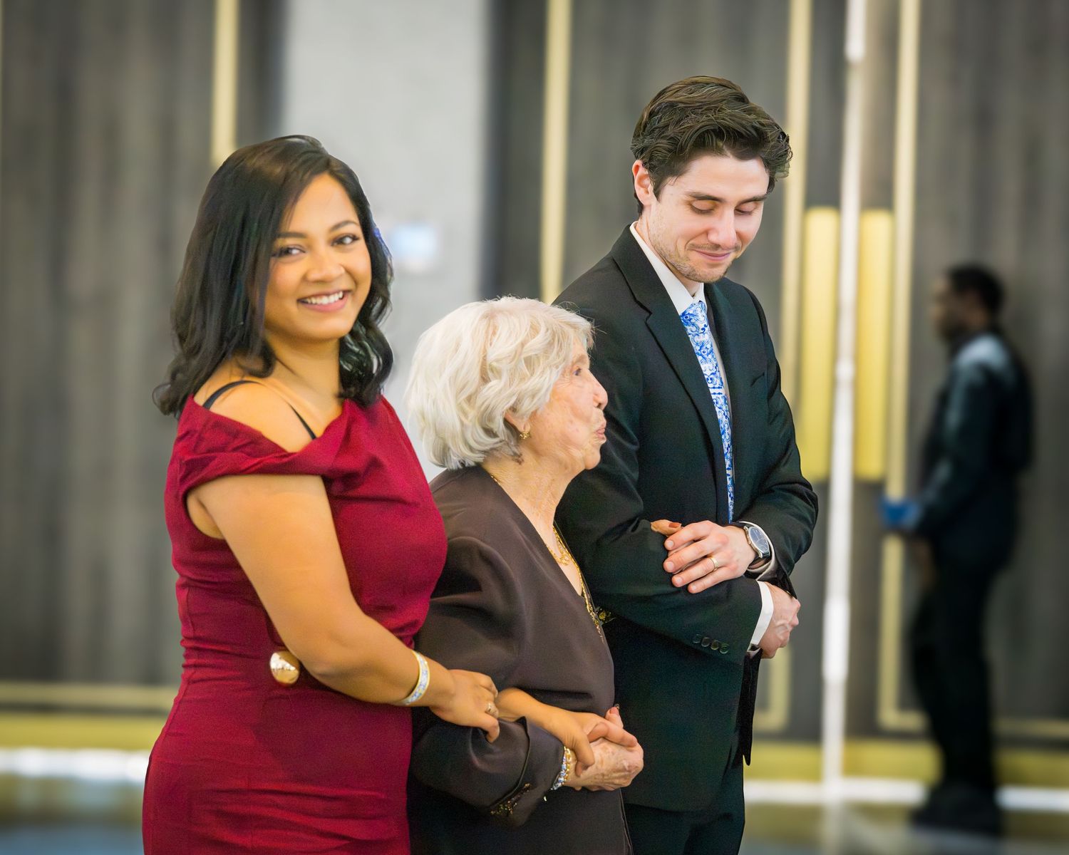 Woman in red dress and young man in suit escort elderly woman at elegant formal event.