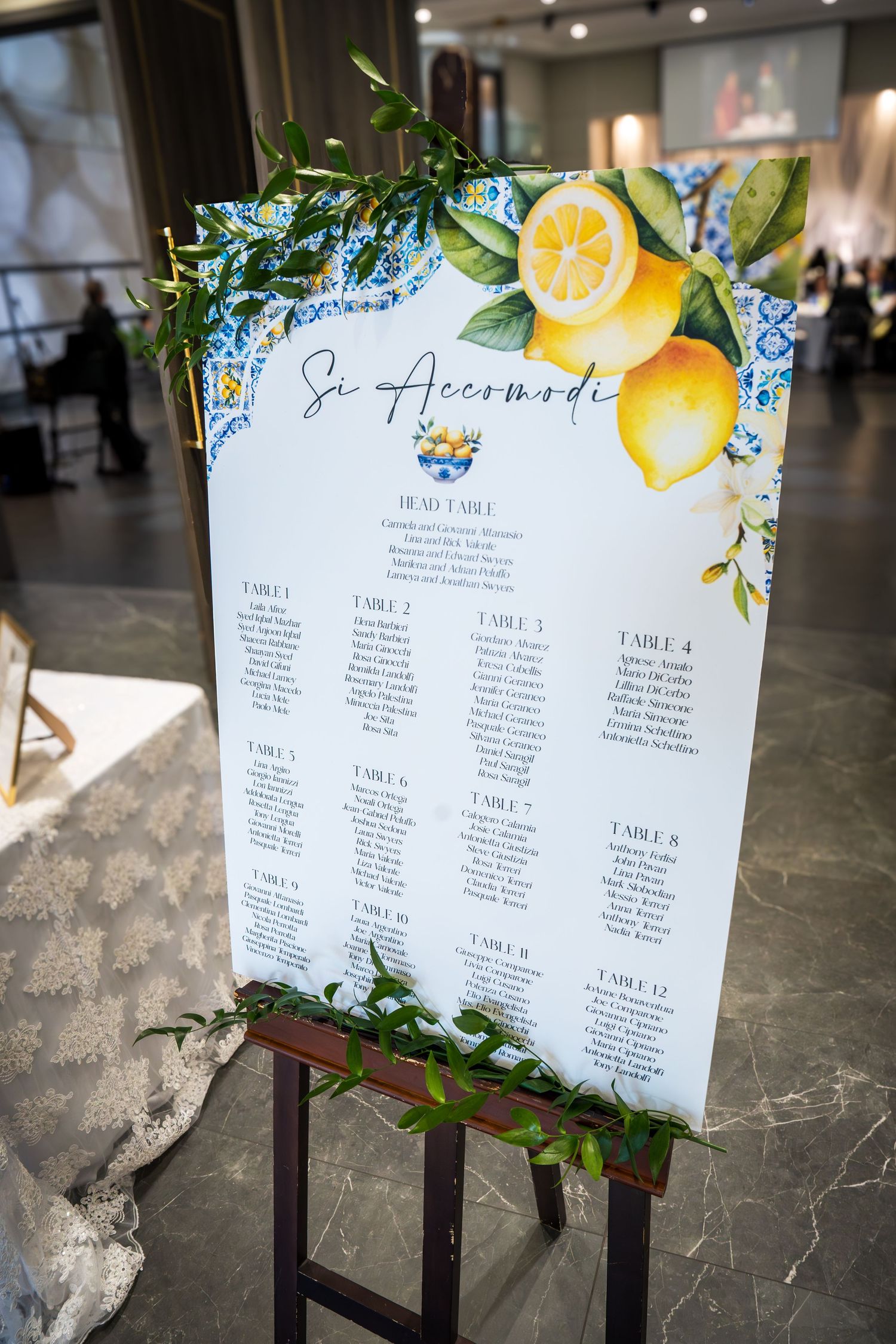 Wedding seating chart sign reading Si Accomodi decorated with lemons and blue tiles on a wooden easel.