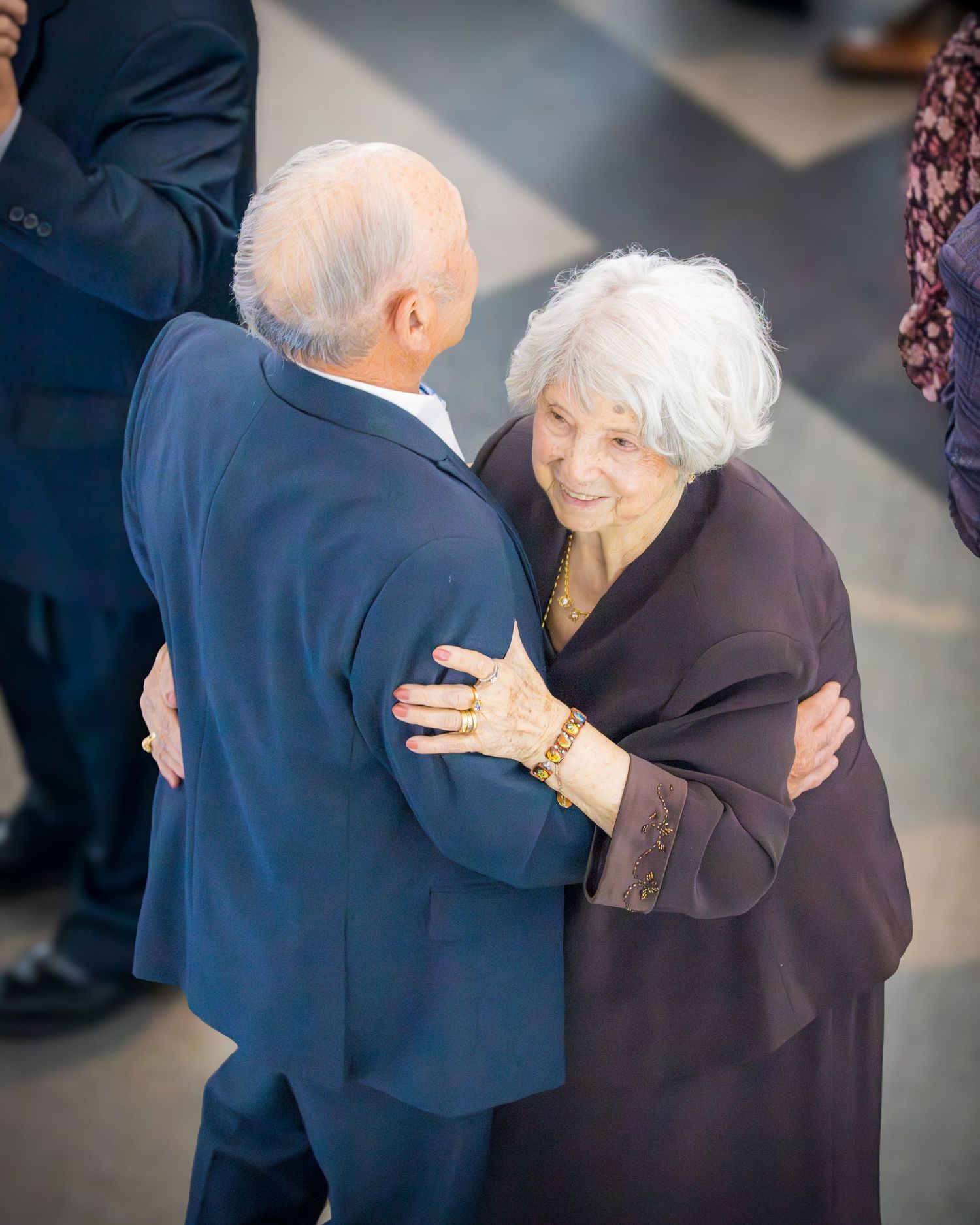 Elderly woman in purple dress warmly embracing an older man in a blue suit at a formal celebration.