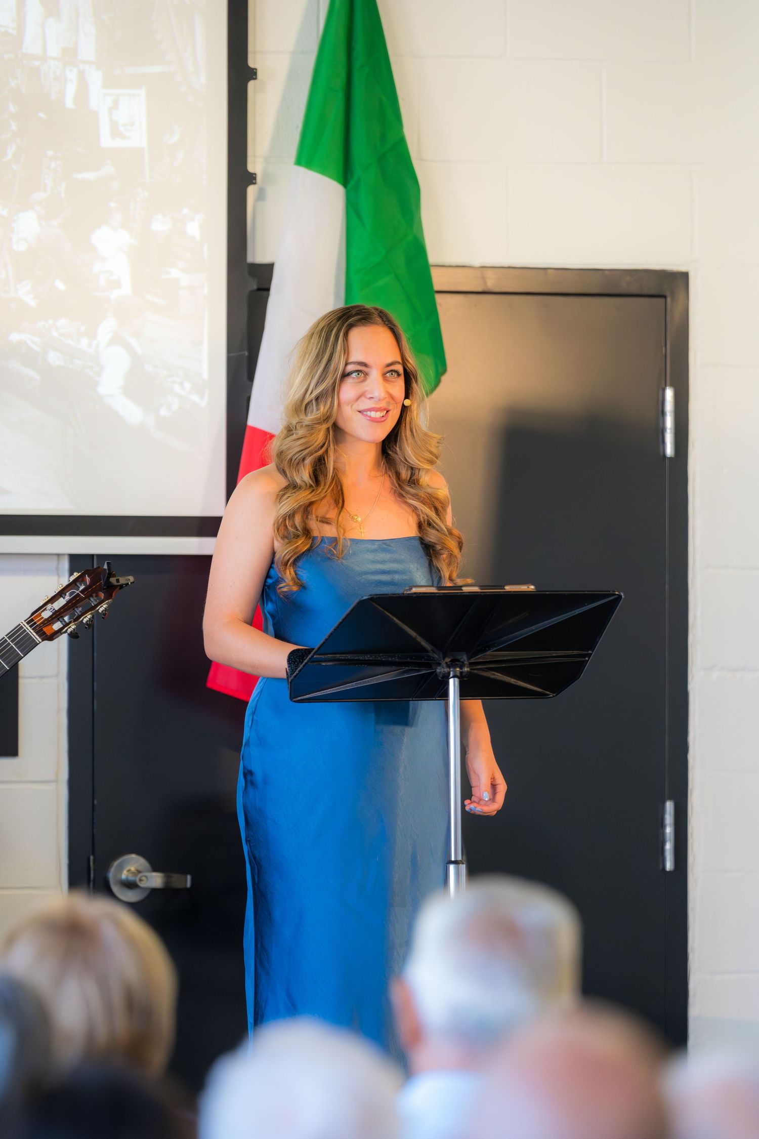 Woman in blue dress performing at podium with Italian flag backdrop.