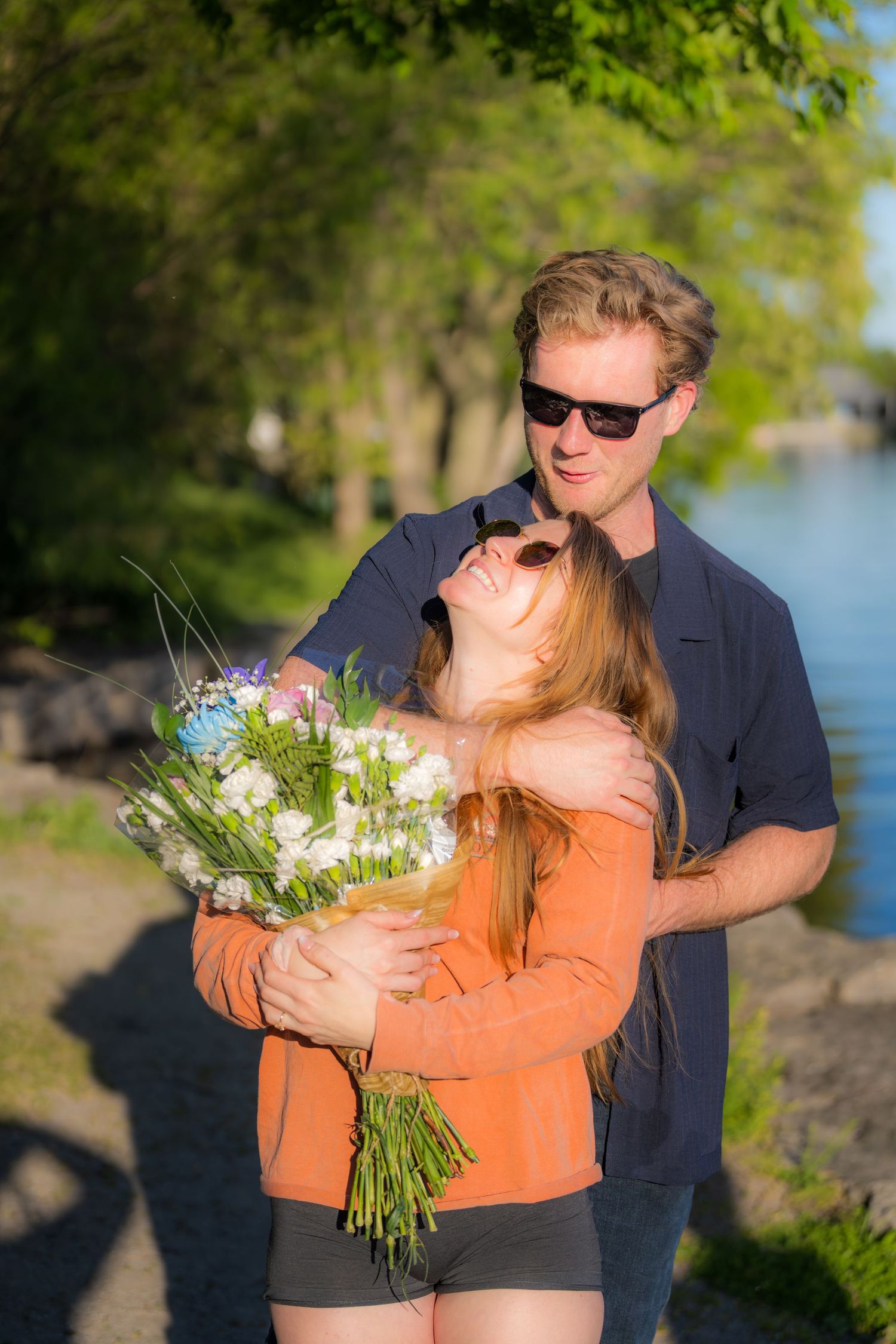 Couple embracing outdoors near a lake, man in sunglasses holding woman with a bouquet of white flowers on a sunny day.