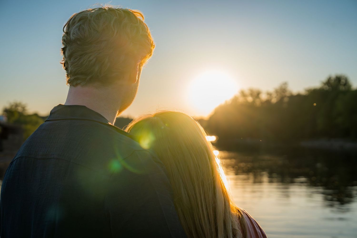 Couple embracing while watching a stunning sunset over a tranquil river surrounded by trees.