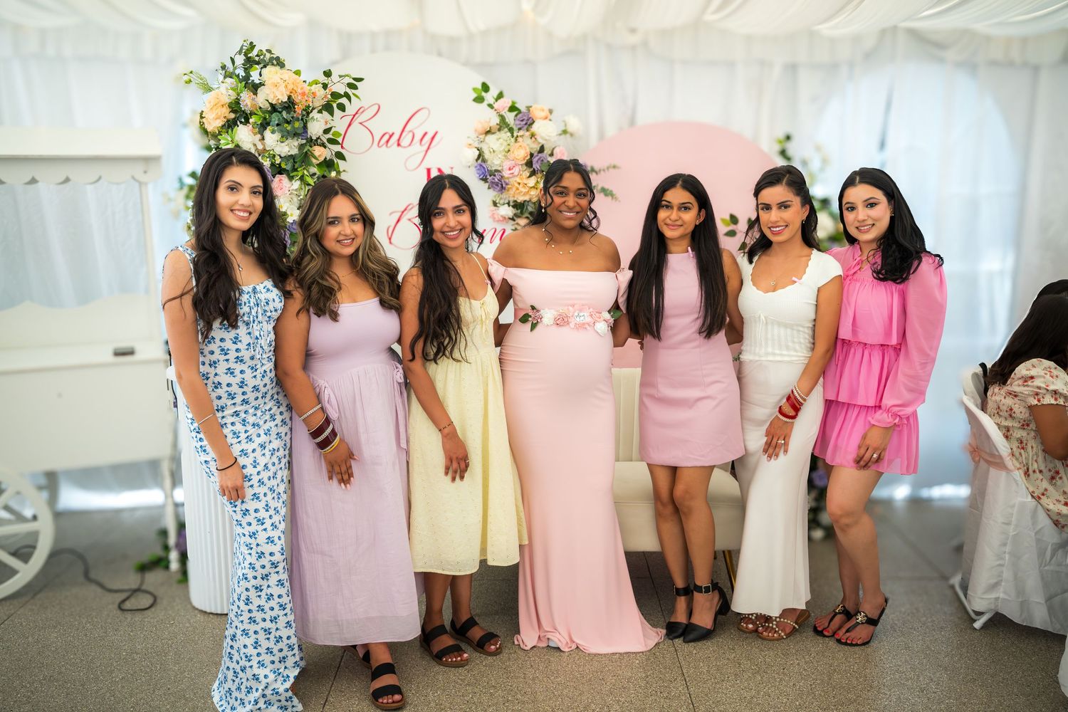 Baby shower guests posing in front of floral wall wearing coordinated pink and pastel dresses.