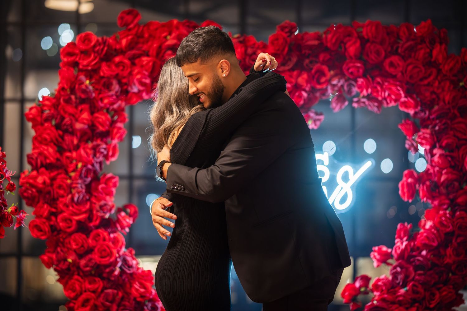 Emotional embrace during proposal with red rose wall and romantic lighting.