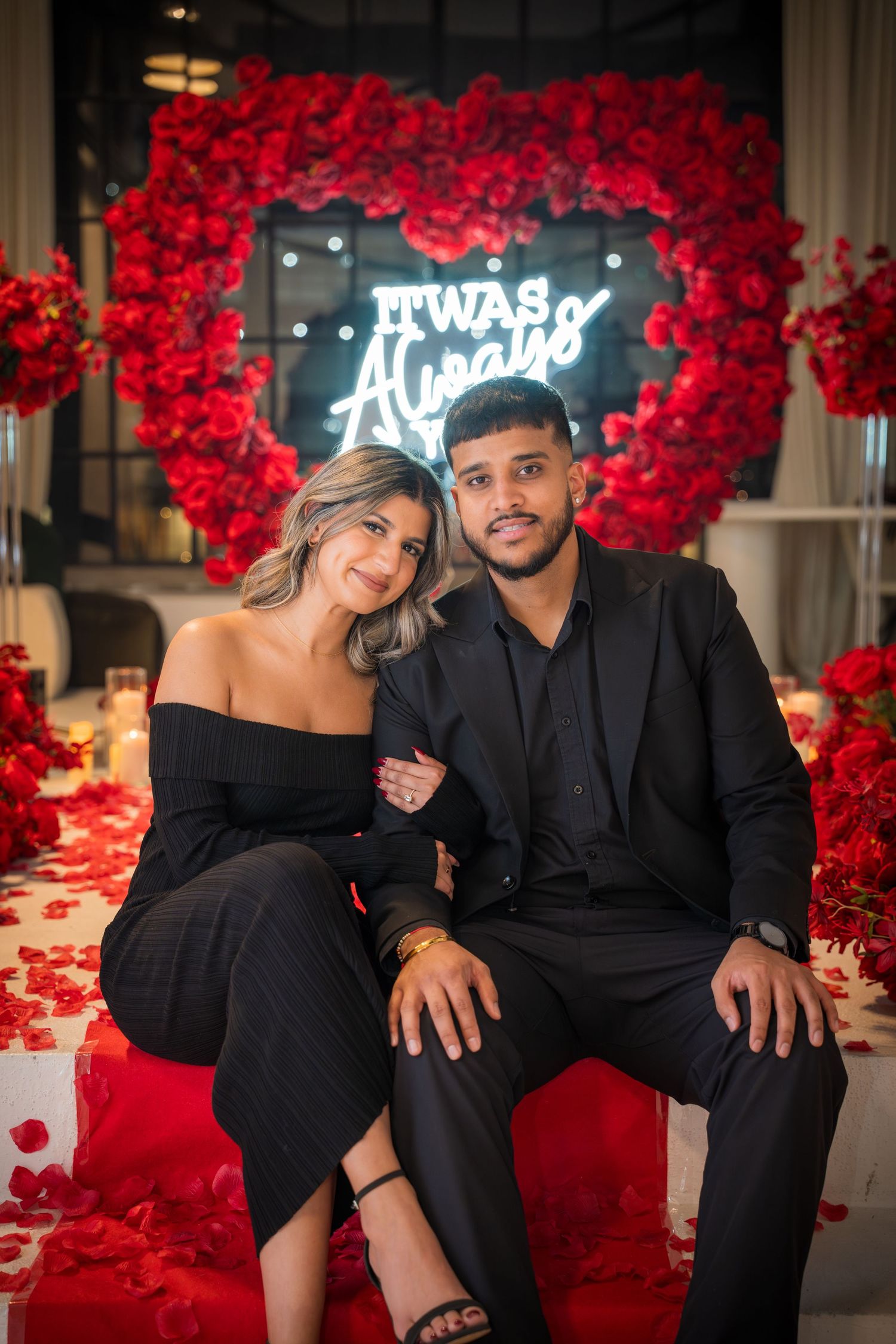 Couple in black outfits sits together on white bench with dramatic red rose backdrop.