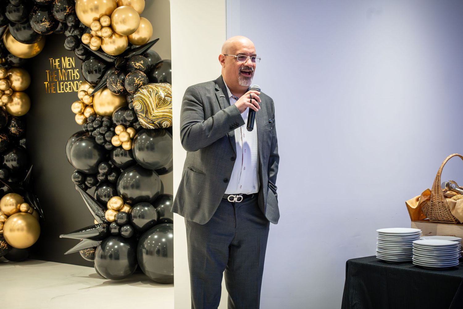 A speaker in a grey suit addresses the audience at a corporate event with gold and black balloon decor.