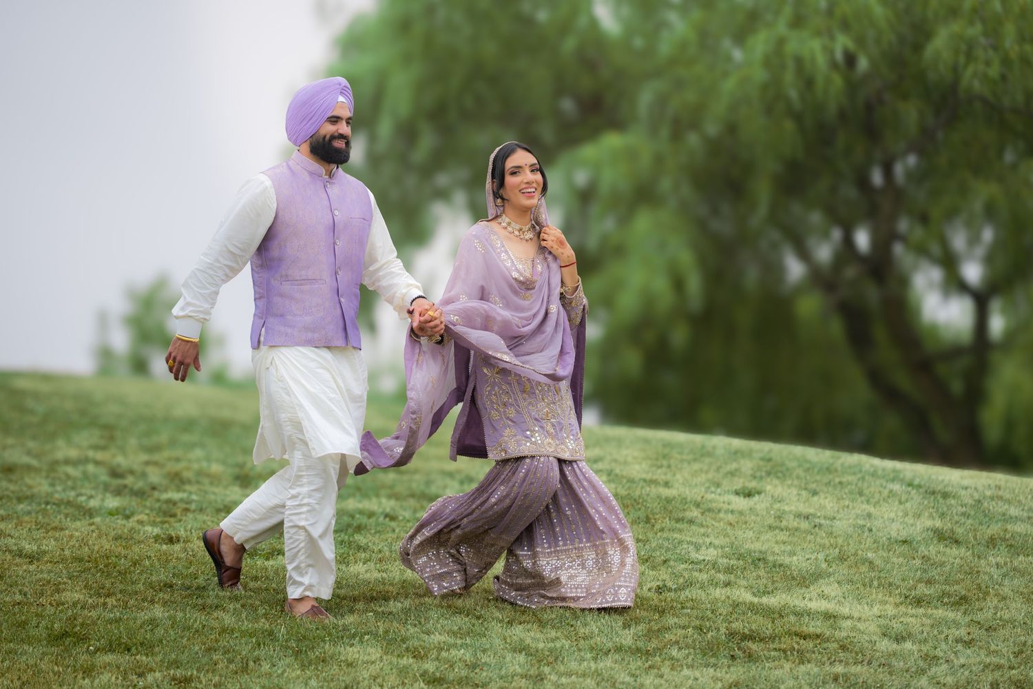 A couple in coordinated purple and white outfits playfully run up a grassy hill in a romantic photoshoot.