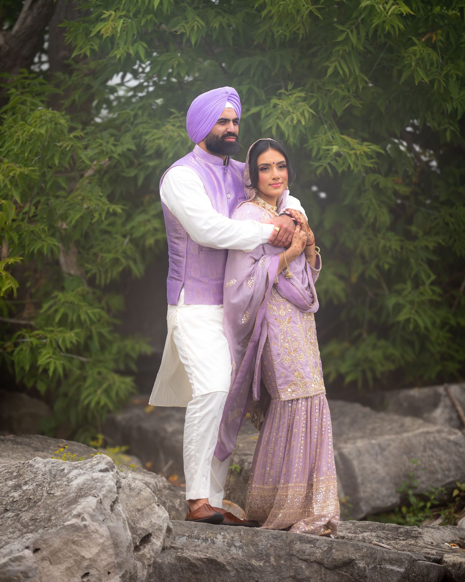 Romantic couple poses together on rocky terrain surrounded by green foliage.