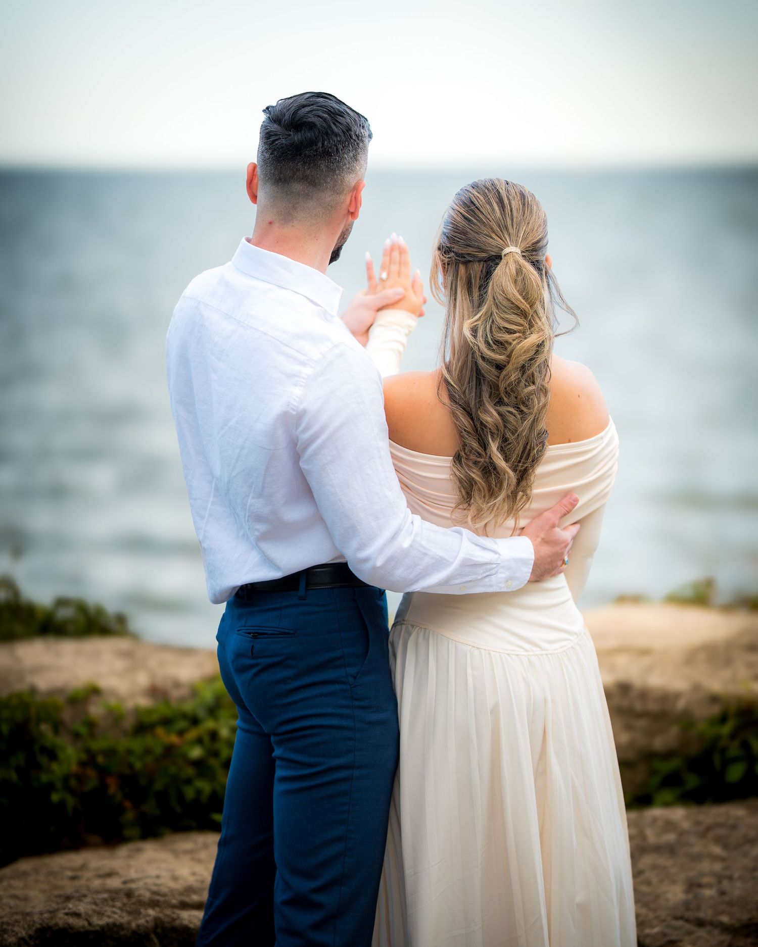 A couple embraces while standing on rocky coastal terrain.