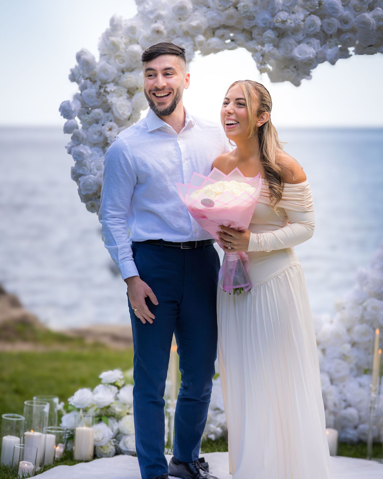 Wedding couple sharing a romantic moment under an elegant white floral arch by the seaside.