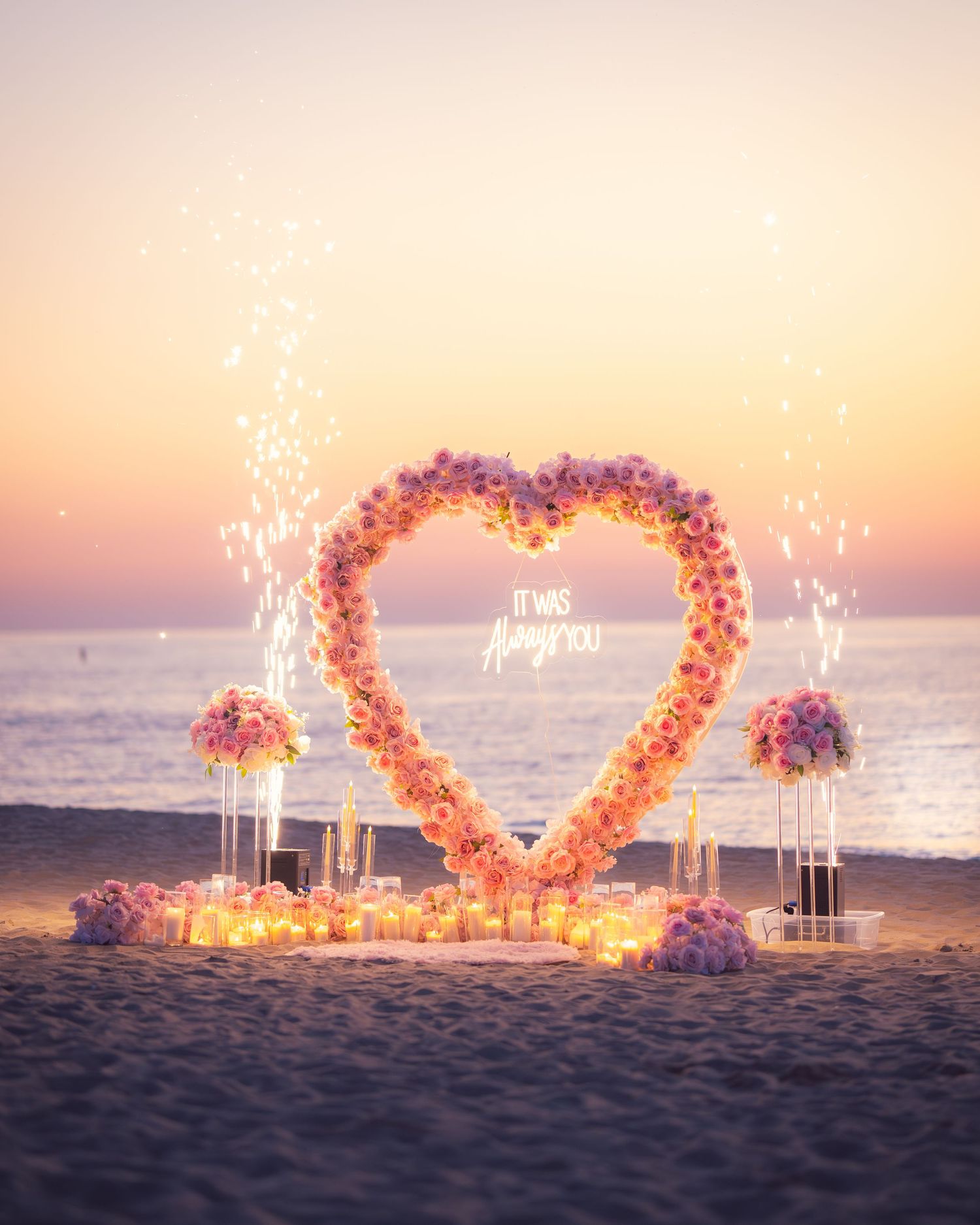 Pink rose heart arch with glowing candles on beach during sunset proposal setup.
