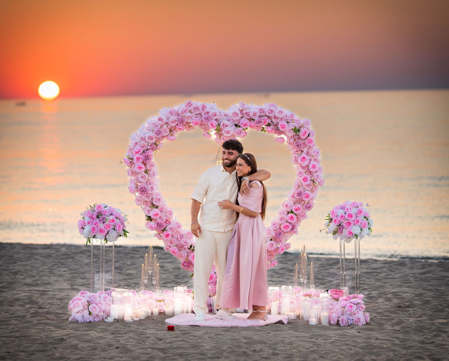 Pink rose-covered heart arch frames a beautiful beach marriage proposal scene during golden hour.