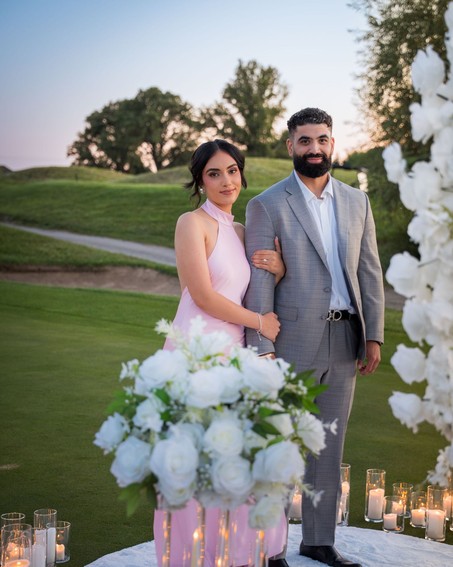 A couple poses together at sunset on a golf course with white floral decorations and glowing candles.
