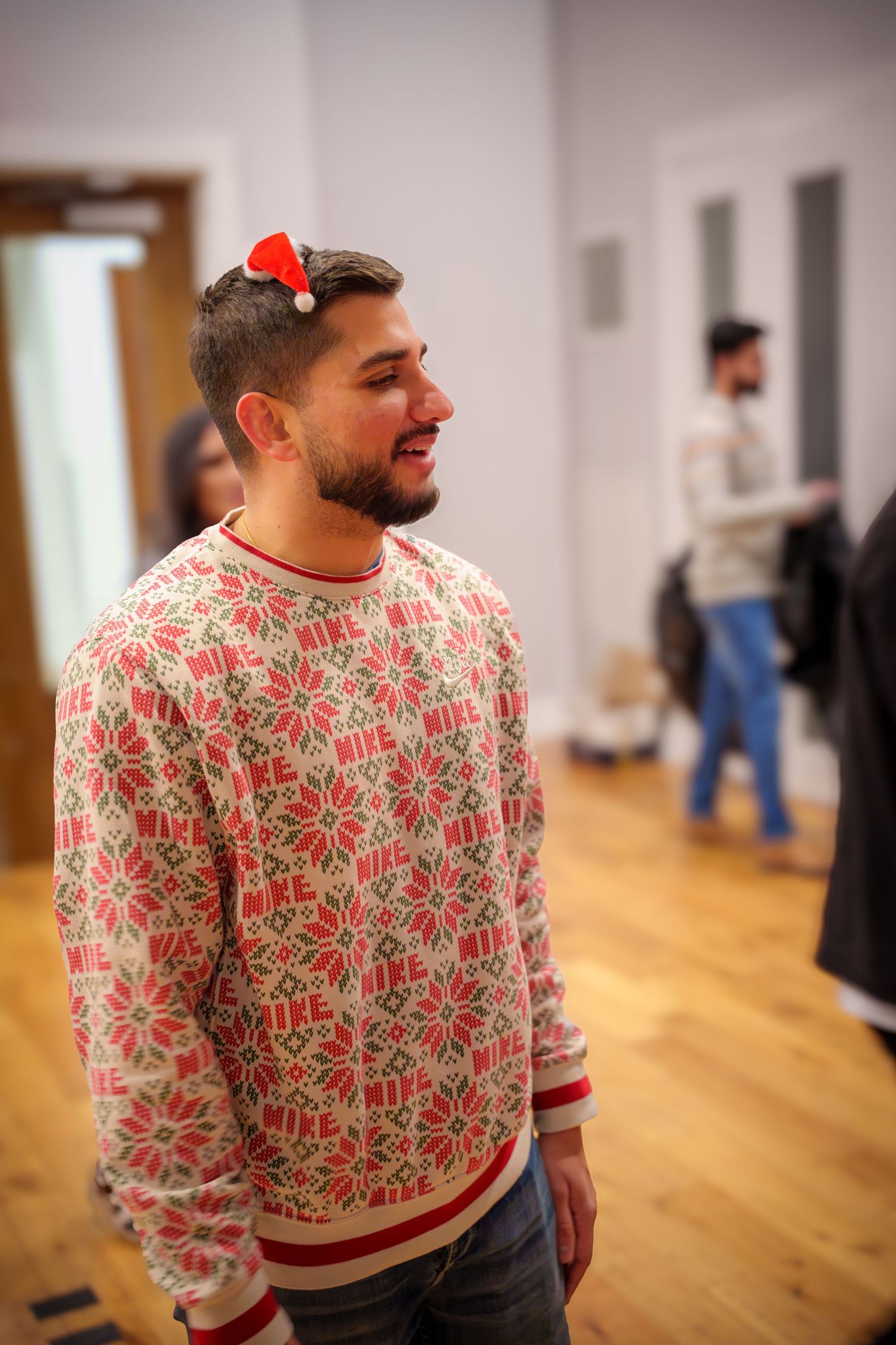 Person wearing a festive red and white patterned sweater smiles while standing in a room with wooden floors.