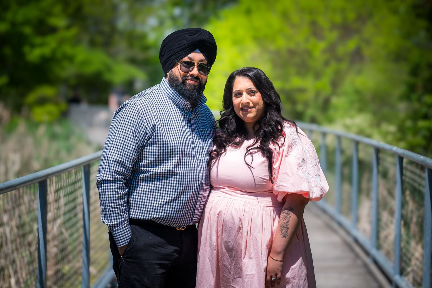 A couple poses on a bridge surrounded by lush greenery, the woman in a pink maternity dress.