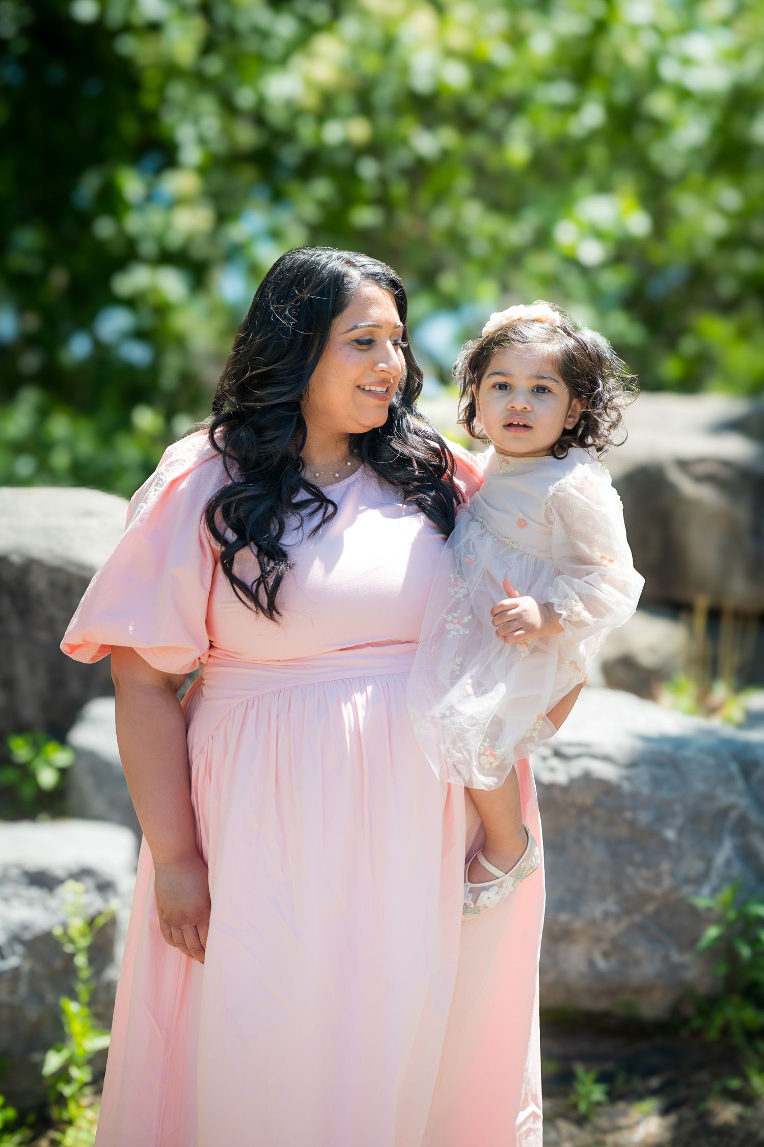 Mother in pink maxi dress holding young toddler girl in white dress outdoors.