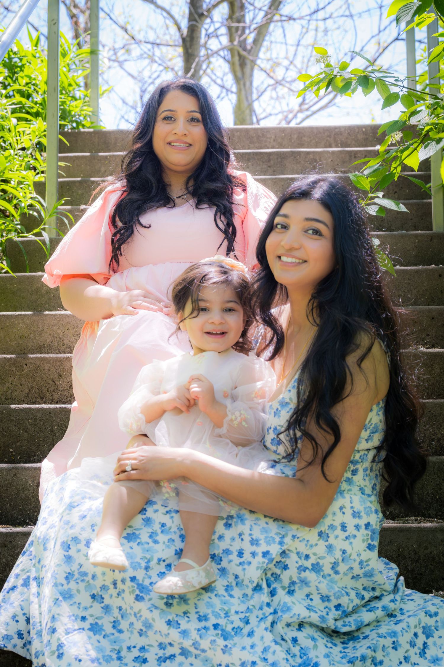 Two women in floral and pink dresses sit on outdoor steps with a smiling baby girl.
