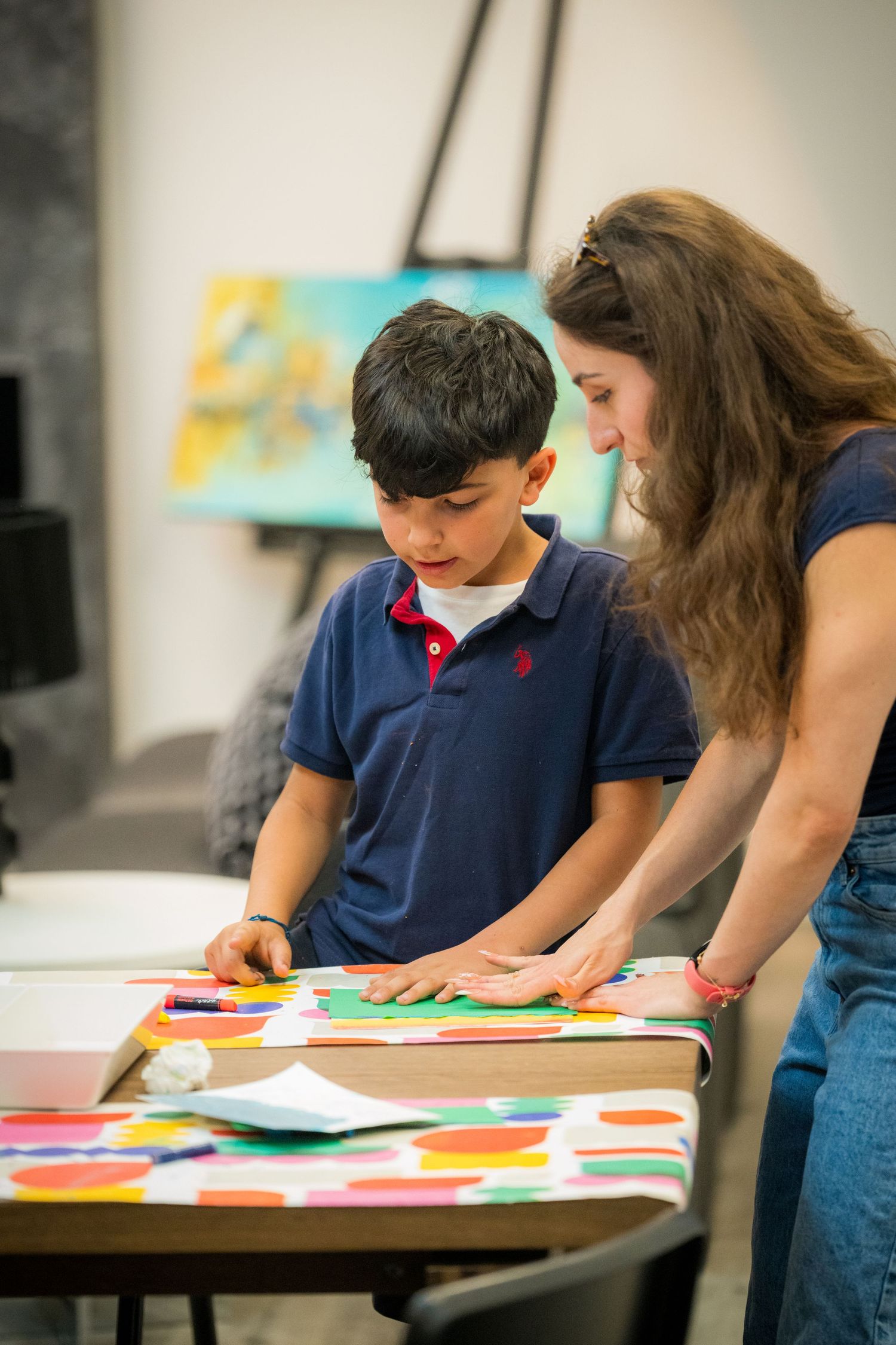 A woman helps a young boy with a colorful paper craft project at an art table.