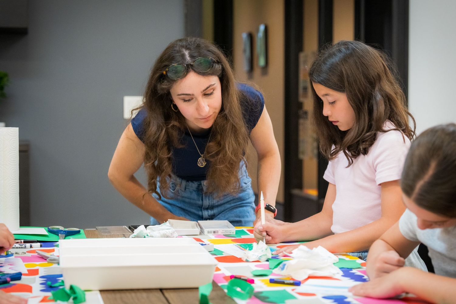 A female art instructor leans over to help a girl with a colorful paper collage project.