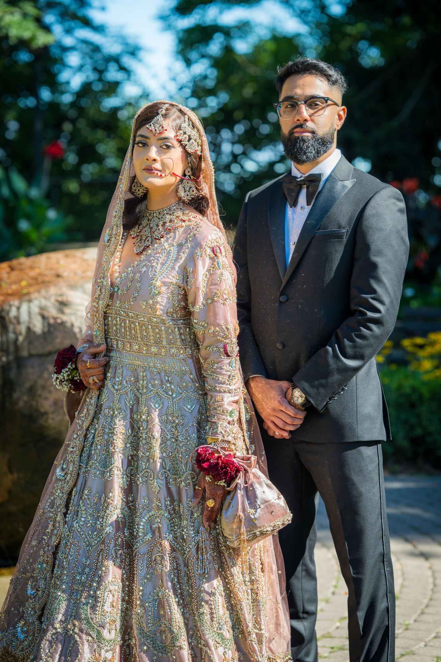 South Asian wedding couple in traditional attire standing on garden path with floral decorations.