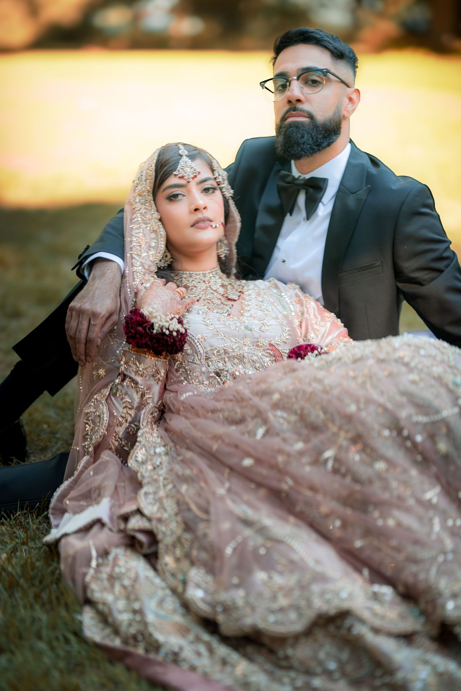 Romantic wedding portrait of couple in traditional South Asian attire relaxing on grass at sunset.