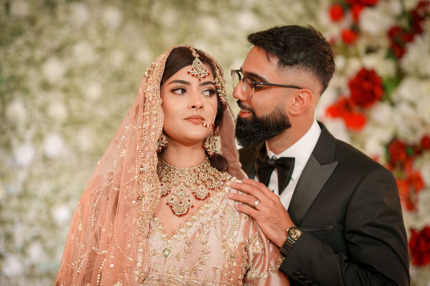Couple in traditional South Asian wedding attire pose against floral backdrop with red roses.