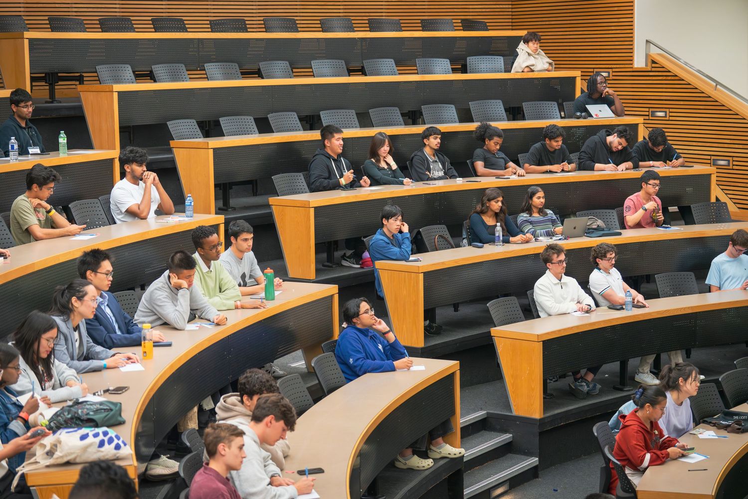 Wide view of a lecture hall showing multiple rows of students seated at long desks with wood-paneled walls.