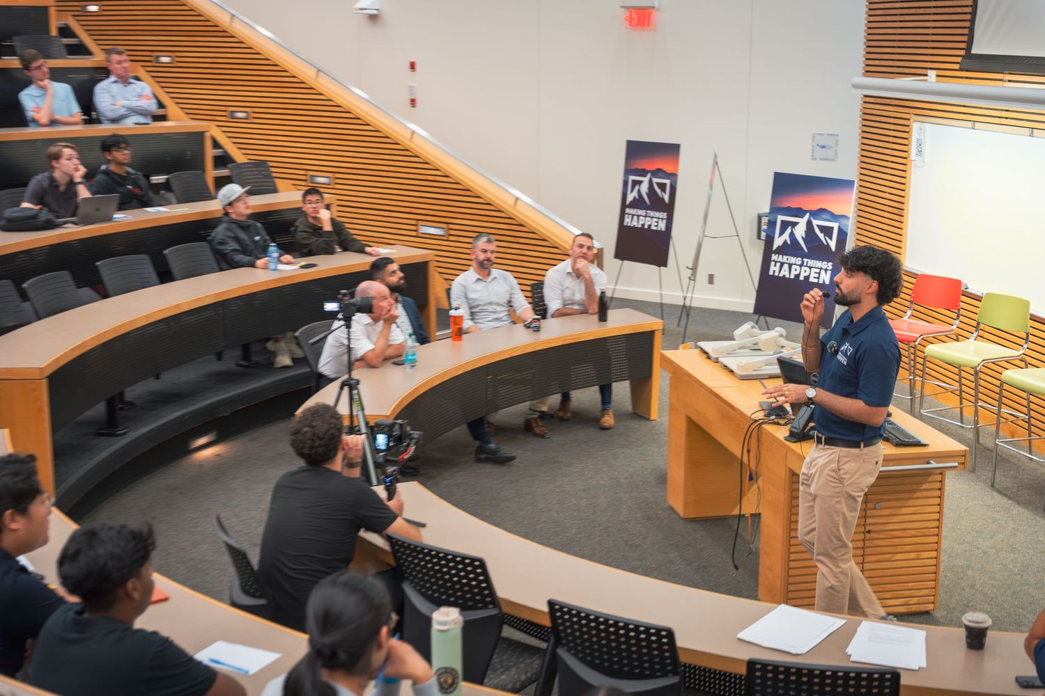 Students sit in a curved lecture hall with wooden accents while attending an academic presentation.