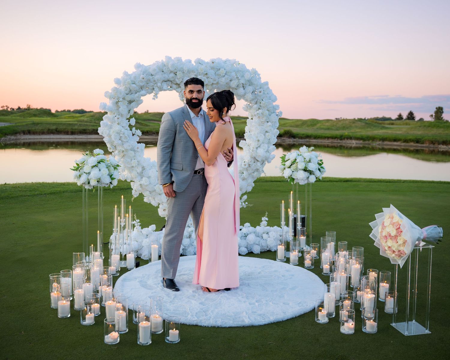 Romantic proposal setup at sunset with white floral arch, candles, and sparklers on a golf course.