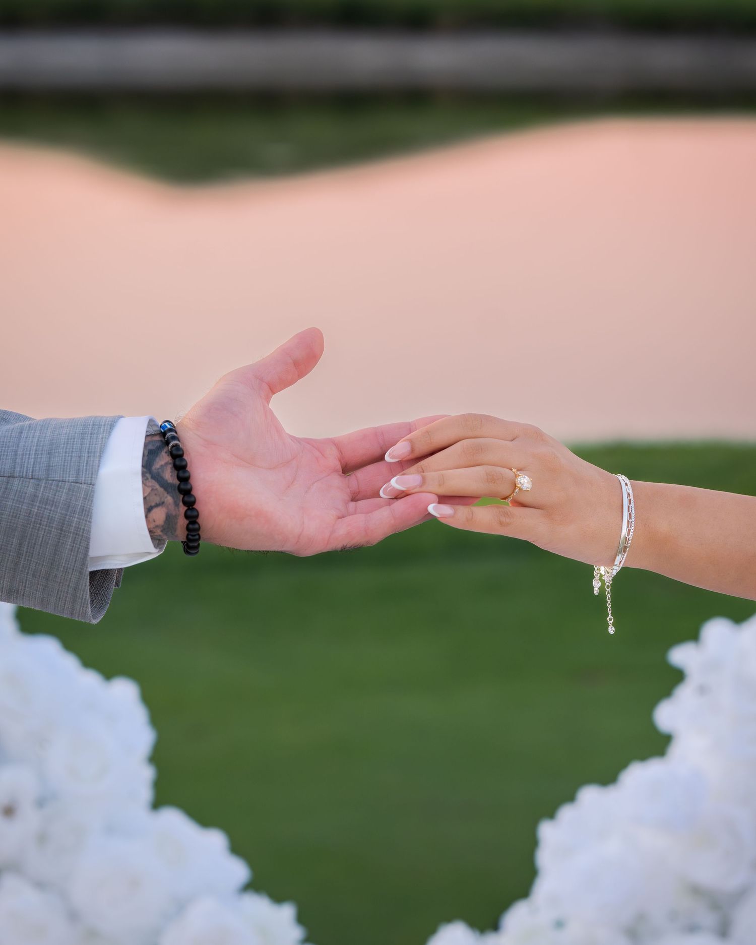 Hands reaching towards each other with white floral decorations and pink sunset in the background.