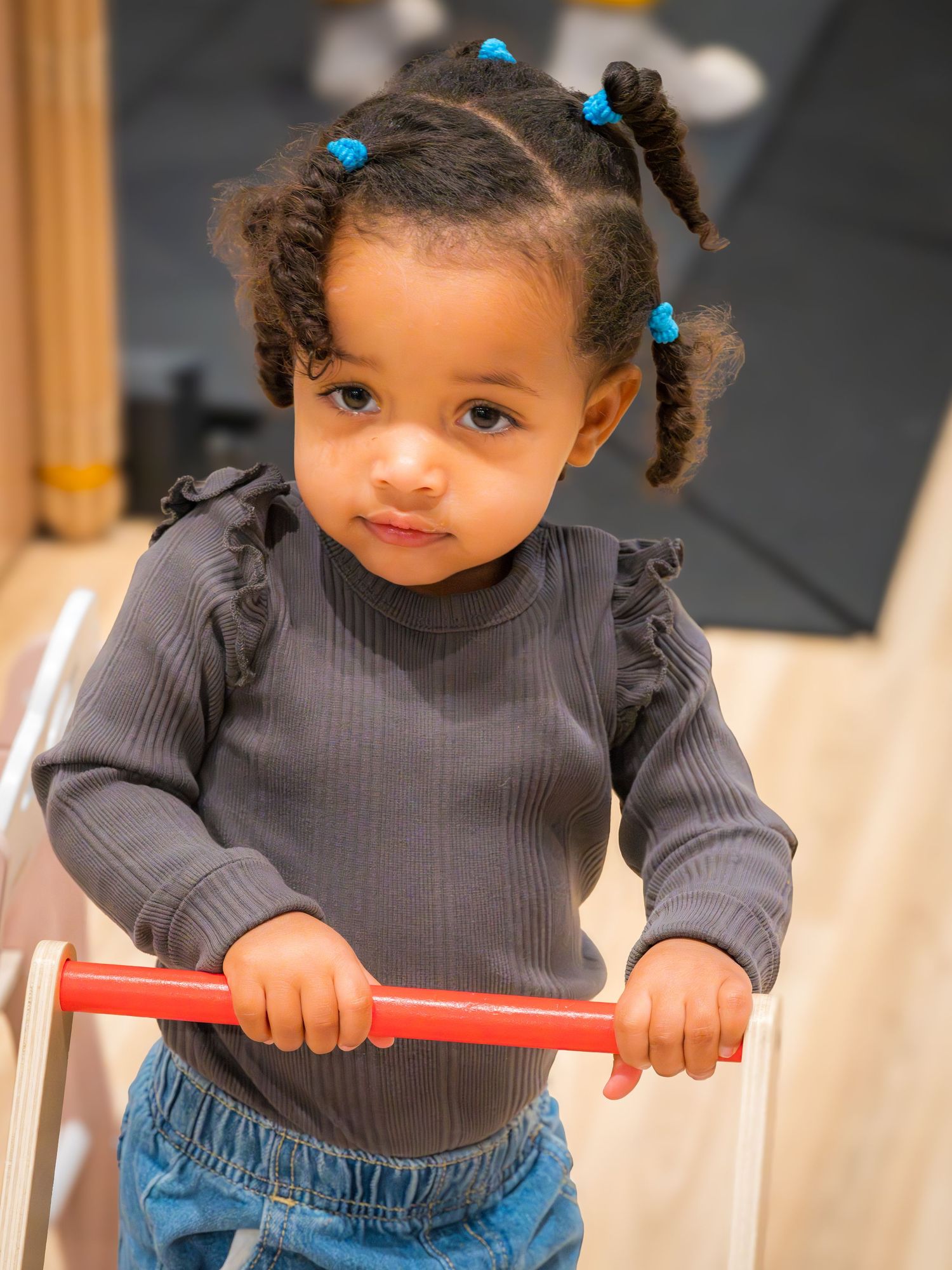 An adorable toddler girl with blue hair ties holds a red bar while standing in a play area.