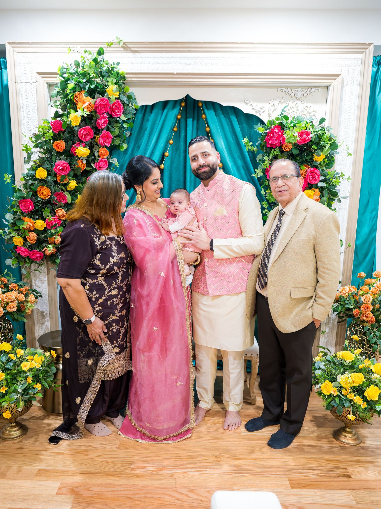 Smiling family posing with baby girl in pink lehenga at colorful floral Lohri celebration backdrop.