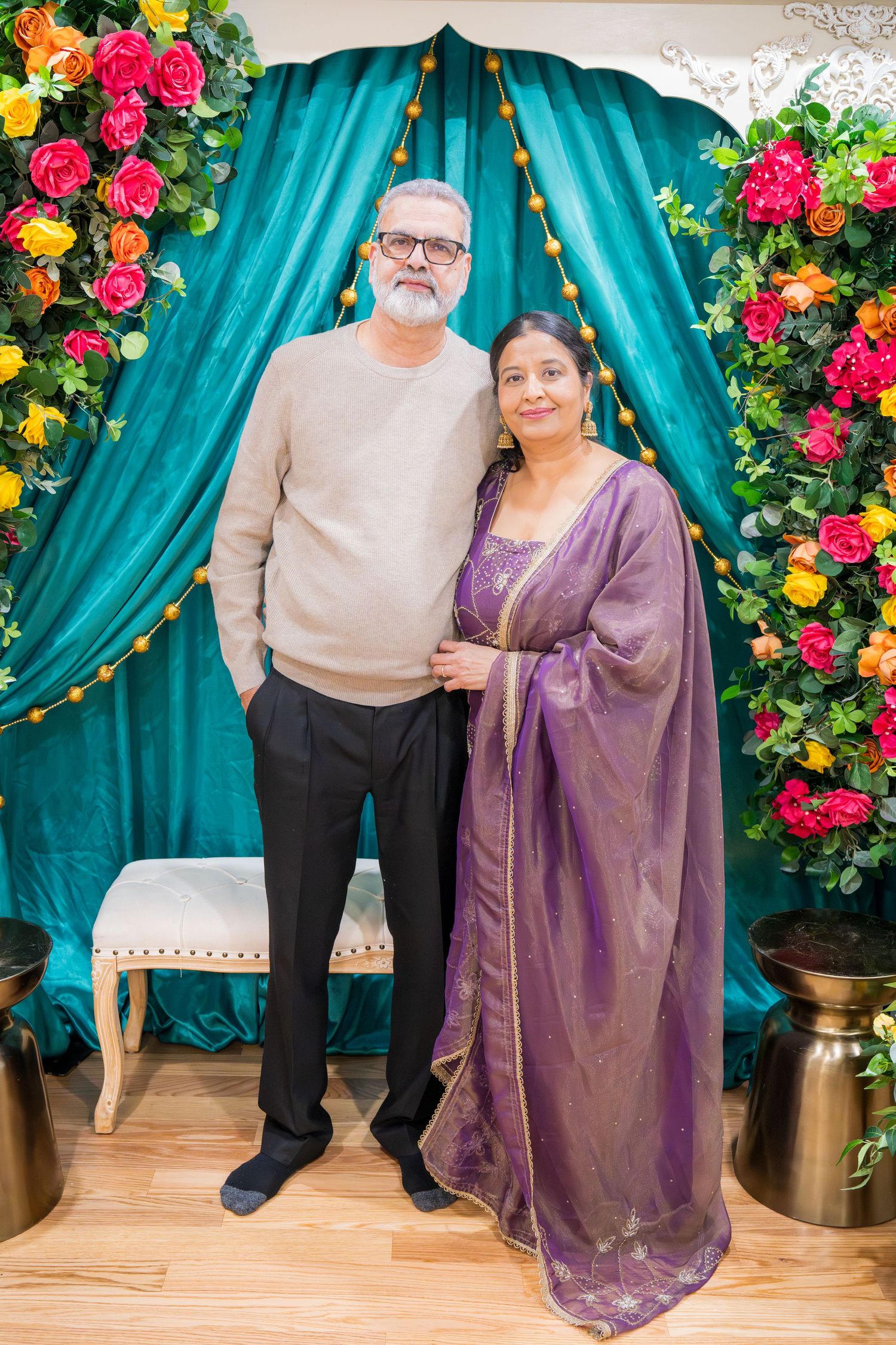 Older couple posing together, man in beige sweater and woman in purple saree at floral backdrop.