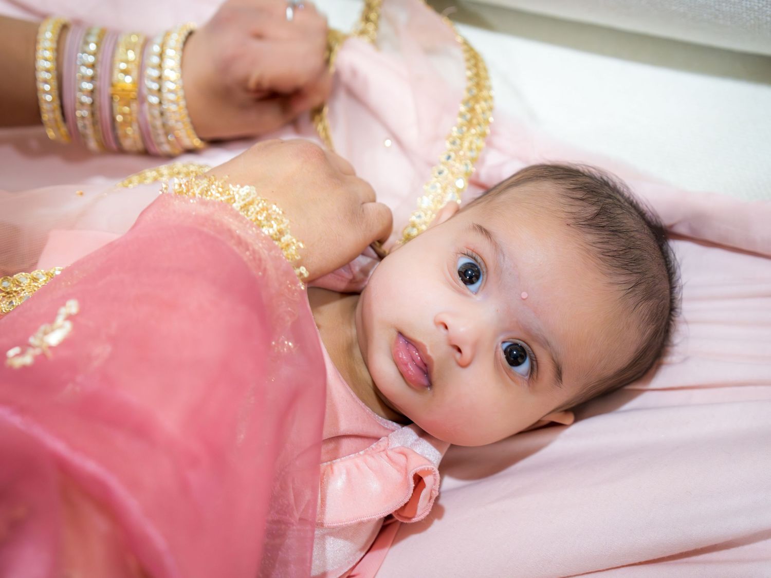 Close-up of an adorable baby in a pink embroidered outfit with a bindi, held by a woman wearing gold bangles.