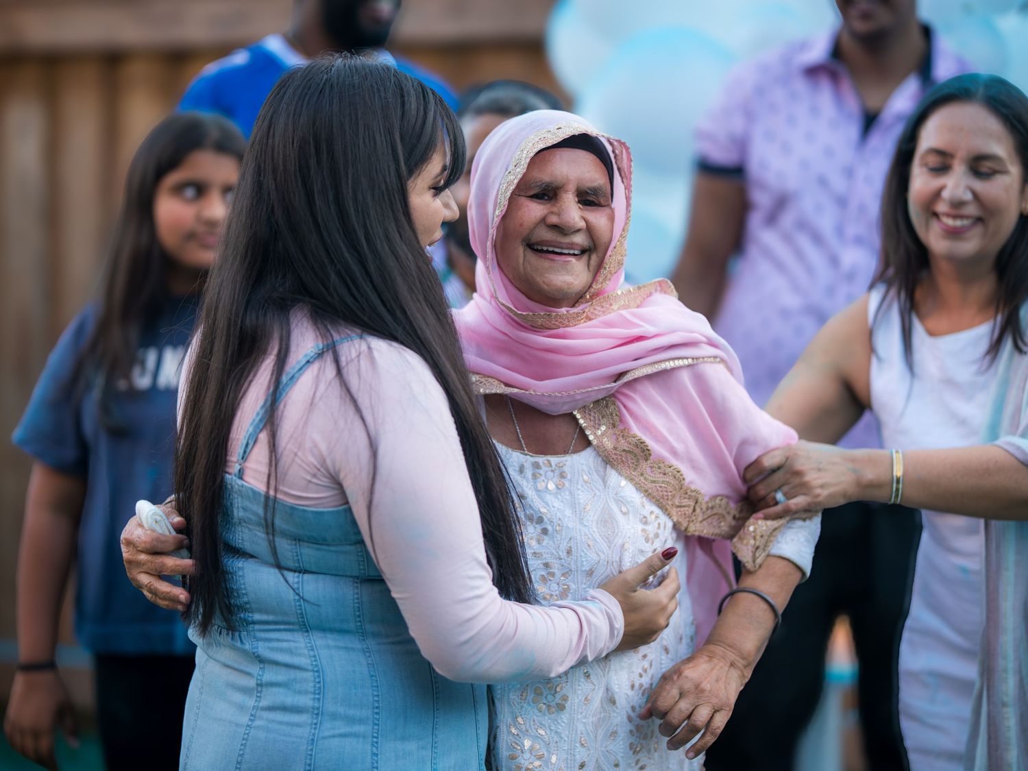 Women in colorful traditional South Asian attire laughing and embracing joyfully at an outdoor celebration.